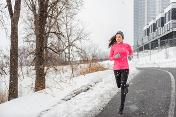 Wall Mural - Winter running Asian girl wearing cold weather clothing for outside exercise in snow storm snowfall during winter training outdoors in city street. Fitness woman exercising.