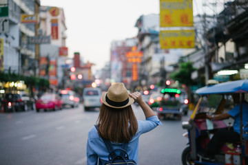 Wall Mural - Young asian woman traveler with blue backpack and hipster hat looking night view on road with tuk tuk Thailand background at China Town Bangkok. Traveling in Bangkok Thailand