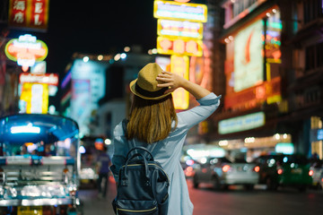 Young asian woman traveler with blue backpack and hipster hat looking night view on road with tuk tuk Thailand background at China Town Bangkok. Traveling in Bangkok Thailand