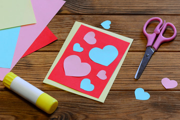 Wall Mural - Valentines day greeting card with paper hearts, scissors, glue stick, paper sheets on a wooden background. Creative Valentines day card design. Project idea using a colored paper. Closeup