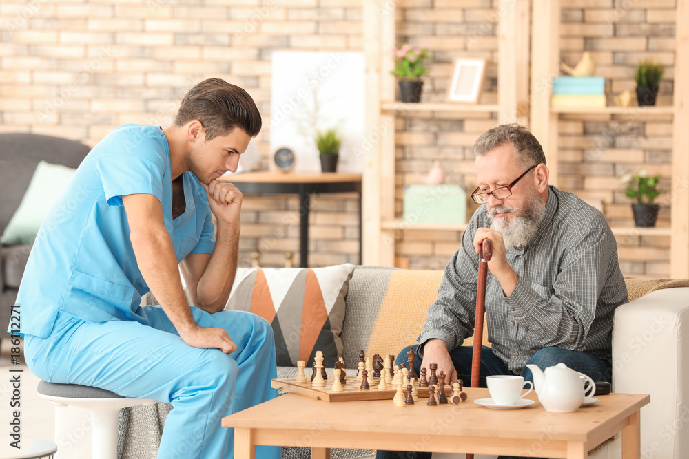 Young caregiver and senior man playing chess at home