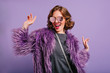 © Look! - Stunning cute girl with curly brown hair posing with pleasure on purple background. Indoor photo of dancing young lady in trendy fur coat laughing to camera.