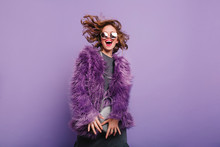 Gorgeous Short-haired Girl In Sunglasses Dancing On Purple Background With Happy Smile. Laughing Female Model In Elegant Fur Coat Posing In Studio With Little Handbag.