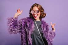 Stunning Cute Girl With Curly Brown Hair Posing With Pleasure On Purple Background. Indoor Photo Of Dancing Young Lady In Trendy Fur Coat Laughing To Camera.