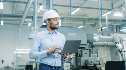 chief engineer in the hard hat walks through light modern factory while holding laptop. successful, 