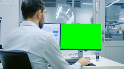 Engineer/ Technician Working on a Personal Computer with Two Displays, One Has a Green Screen Chroma Key Template Great for Mockup. Out of the Office Window Components Manufacturing Factory is Seen.