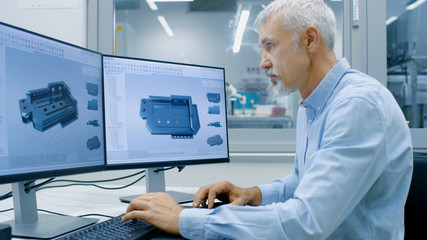 Engineer Working on a Personal Computer with Two Displays, He's Designing New Essential Component in CAD Program. Out of the Office Window Components Manufacturing Factory is Visible.