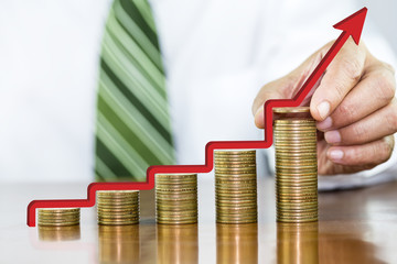 Businessman hand holding one coin and red arrow step up on money coin stack arranged as a graph on wooden table, concept of money growth and saving money