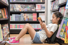 Children's Education Lifestyle With School Girl Kid Reading Book  Sitting On The Floor Of Bookstore Or Library Reading Cartoon Paperback Among  Bookshelf Aisle
