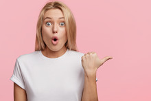 Horizontal Shot Of Stupefied Female Model With Surprised Expression, Keeps Mouth Opened, Not Expect To See Something, Indicates With Thumb At Blank Studio Wall For Your Advertisment Or Promotion