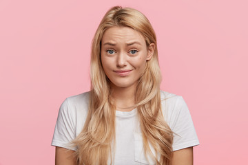 Wall Mural - Adorable blonde young woman without make up, looks with displeased expression, raises eyebrows, can`t understand something, isolated over pink background. People and facial expressions concept