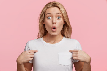 Wall Mural - Portrait of amazed blonde bug eyed female looks with stupefied expression at camera, indicates at blank t shirt space, being shocked with low price, isolated over pink background. Advertising concept