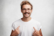 © wayhome.studio - Smiling young male indicates at blank t shirt, has happy expression, glad to advertize new clothes, isolated over concrete background. Unshaven cheerful hipster points at new outfit. Design, clothing