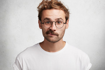 Wall Mural - Isolated shot of indignant bearded male curves lips, looks in bewilderment, wears casual t shirt and square spectacles, isolated over white concrete wall. People, facial expressions, reaction concept