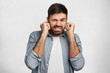 © WHstudio Leushin N - Handsome bearded young male model plugs ears with displeased expression as hears annoying loud noise or roaring not far from him, isolated over white concrete wall. Not pleasant irritated sound