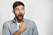 Horizontal portrait of shocked young male has thick beard and mustache looks with widely opened mouth and bugged eyes aside, points with fore finger at blank copy space for your advertisment