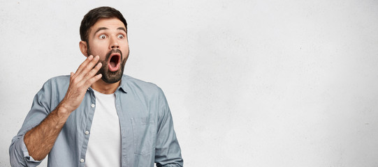 Wall Mural - Horizontal studio shot of shocked frightened male model has hush reaction as sees his phobia, keeps mouth opened, dressed casually, isolated over white background with copy space for your text