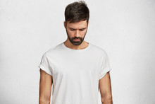 Handsome Brunet Male With Trendy Hairdo, Has Muscular Body, Wears Caual White T Shirt, Looks Down As Notices Something On Floor, Poses Against Concrete Background. Attractive Man Hangs Head.