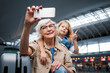 © Yakobchuk Olena - Lovely memories. Portrait of happy gray-haired granny is posing at international airport with her cheerful cute granddaughter. She is holding smartphone while looking at screen and taking photo