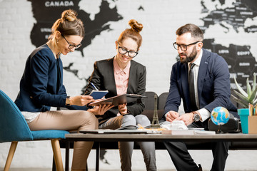 Wall Mural - Young businesscouple choosing a trip with agent sitting at the travel agency office with world map on the background