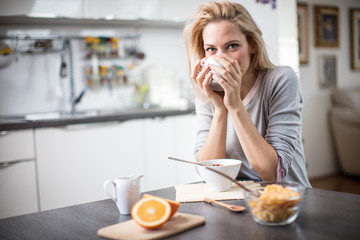 Wall Mural - Beautiful blond  caucasian woman posing in her kitchen, while drinking coffee or tea and eating a healthy breakfast meal full of cereal and other healthy foods, including fruit