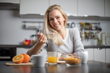 Wall Mural - Beautiful blond  caucasian woman posing in her kitchen, while drinking coffee or tea and eating a healthy breakfast meal full of cereal and other healthy foods, including fruit