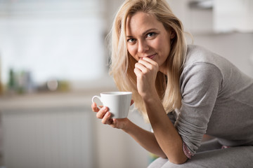 Wall Mural - Beautiful blond  caucasian woman posing in her kitchen, while drinking coffee or tea and eating a healthy breakfast meal full of cereal and other healthy foods, including fruit