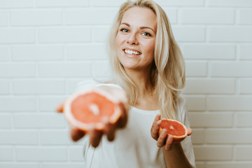 Wall Mural - Beautiful blond  caucasian woman posing in front of a white brick backround and  having fun with a set of two red oranges