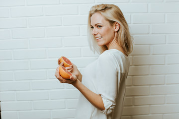 Wall Mural - Beautiful blond  caucasian woman posing in front of a white brick backround and  having fun with a set of two red oranges