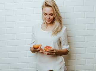 Wall Mural - Beautiful blond  caucasian woman posing in front of a white brick backround and  having fun with a set of two red oranges