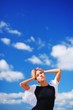 © raisondtre - Portrait of a pensive attractive woman with raised hands against blue sky with floating white clouds, close-up