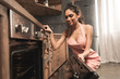 © LIGHTFIELD STUDIOS - sexy young woman in apron opening oven and smiling at kitchen