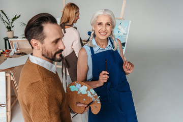 Sticker - cheerful senior woman holding paintbrush and smiling at camera at art class