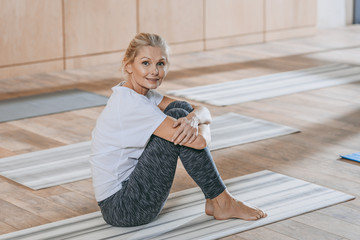 beautiful mature woman sitting on yoga mat and smiling at camera at training class