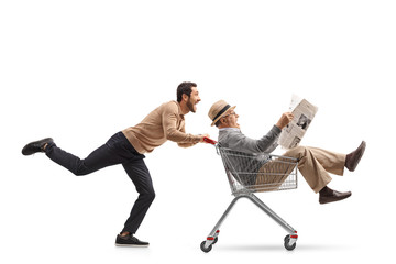 Wall Mural - Mature man with a newspaper riding inside a shopping cart being pushed by a young man