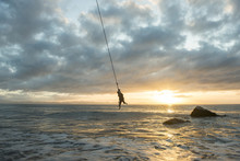Silhouette Man Hanging On Rope Over Sea Against Cloudy Sky During Sunset