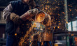 Side close up view of professional focused worker man in uniform working on the metal pipe sculpture with an electric grinder while sparks flying in the industrial fabric workshop.