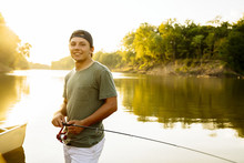 Portrait Of Smiling Young Man With Fishing Rod Standing At Lakeshore Against Clear Sky During Sunset