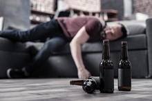 Alcoholic Drink. Selective Focus Of Beer Bottles Standing On The Floor With Bearded Man Sleeping In The Background
