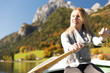 Junge, hübsche Frau rudert mit einem Ruderboot auf einem See in den Bergen Alpen an einem wunderschönen Herbsttag. Sie genießt die Sonne, liest ein buch und macht ein Selfie mit ihrem Handy Kamera