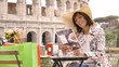 Beautiful young woman tourist reading menu sitting at the table of a bar restaurant in front of the Colosseum in Rome. Elegant dress with large hat and colorful shopping bags on a summer day.