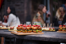 Abstract Blurry Food Background. Small Burgers With Meat, Salads Cheese And Tomatoes On Wooden Plate During Summer Barbecue Party Outdoors. Friends Preparing Food, Chatting And Having Drinks Together