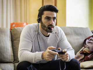 Young handsome man playing video game and talking with online players through headset, while sitting on couch at home at night