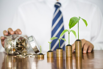 Money growing plant and soil on money coin stack arranged as a graph with businessman pour the mix coins out of the clear bottle on wooden table, concept of money growth and business investment