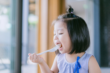 Happy Little asian girl eating sausage and she have fork in her mouth.Eating healthy breakfast in the morning.