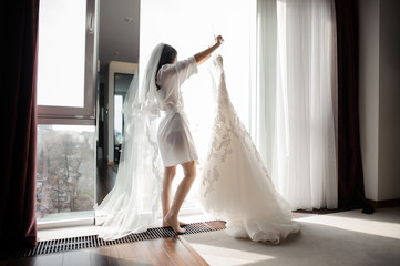 bride in dressing gown and veil holding wedding dress on a hanger