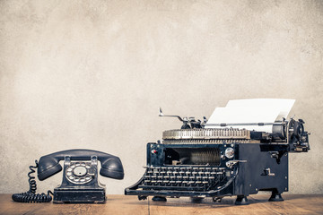 Retro old black telephone and aged typewriter with paper blank on wooden desk front concrete wall background. Vintage style filtered photo