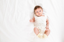 Top View Of Cute Adorable Baby Girl Wearing White Body In Bedroom Looking At Camera. Newborn Child Relaxing And Smiling In The Bed In Children Nursery. Family Morning At Home
