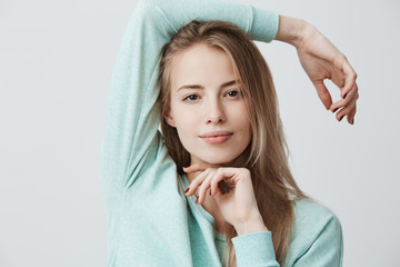 Wall Mural - Headshot of happy pleased blonde girl of European appearance with dark eyes wearing blue long-sleeved top looking and smiling at camera, standing isolated against gray background. Positive emotions