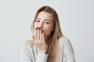 Wall Mural - Pleasant-looking beautiful blonde female with red manicure blows kiss at camera, sends goodbye to boyfriend, poses against gray background with copy space for text or advertisment. Distant kiss